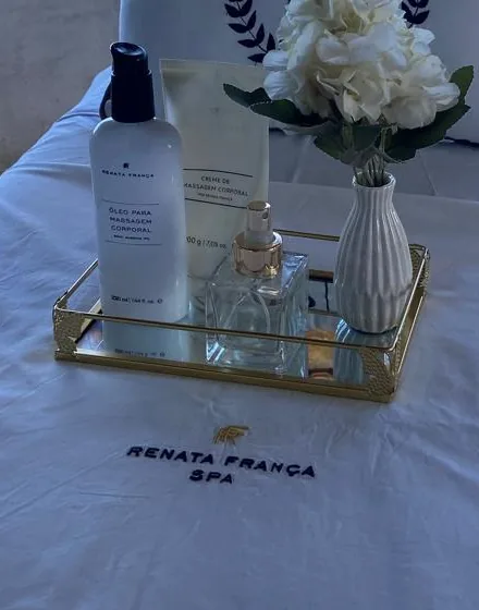 Bandeja dourada com óleos, perfume e flores brancas sobre o lençol bordado Renata França Spa