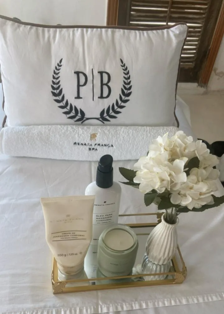 Travesseiro branco bordado com o monograma PB entre louros, com bandeja dourada, flores brancas, vela e óleos do Método Renata França